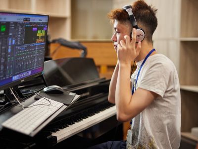 A man adjusting his headphones, sat at a laptop and piano keyboard working on music production software